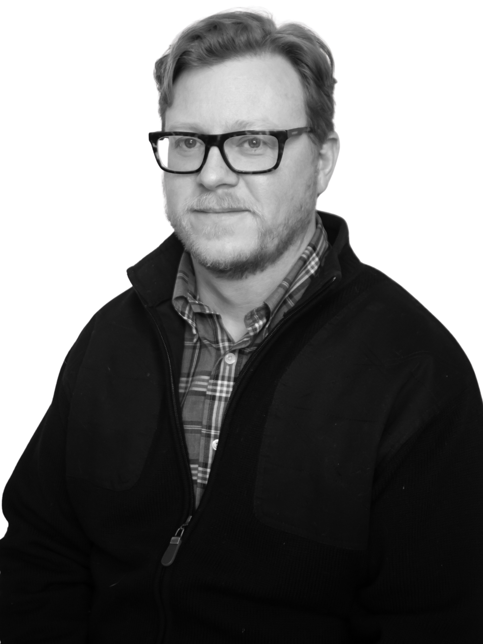 A man with light hair, glasses, and a beard wears a plaid shirt and dark jacket, looking at the camera with a neutral expression. The background is plain and light-colored.
