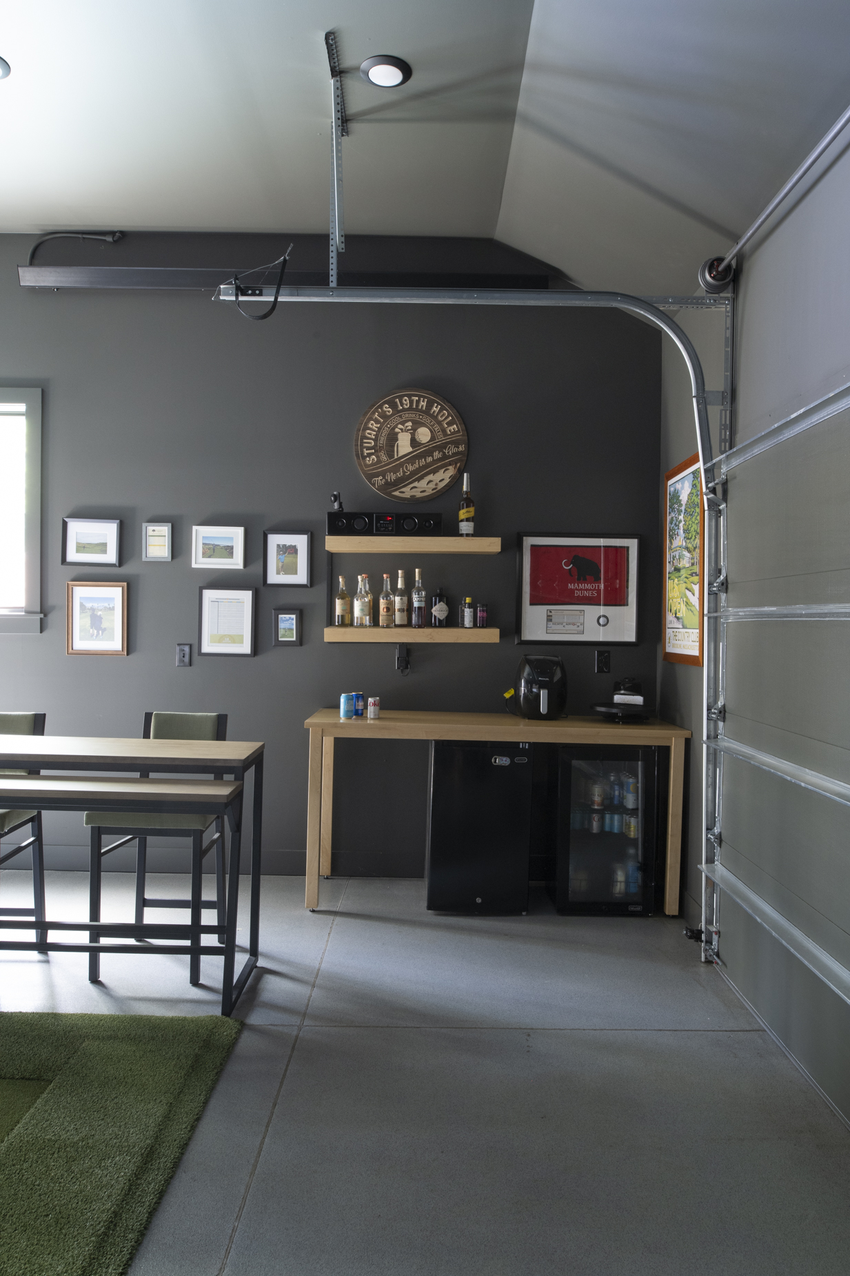 A garage bar in a lake home features dark walls, a wooden table with chairs, framed art, shelves with bottles, a small refrigerator, coffee maker, custom cabinetry and millwork, and an open garage door letting in natural light.