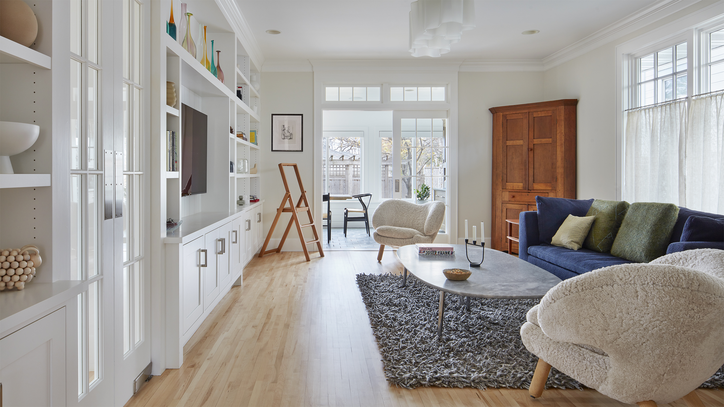 Bright living room with white built-ins, TV, gray rug, marble coffee table, blue sofa, and two fluffy chairs—a true revival of contemporary comfort. Large windows let in natural light, illuminating the wooden cabinet in the corner and a small desk by the window.