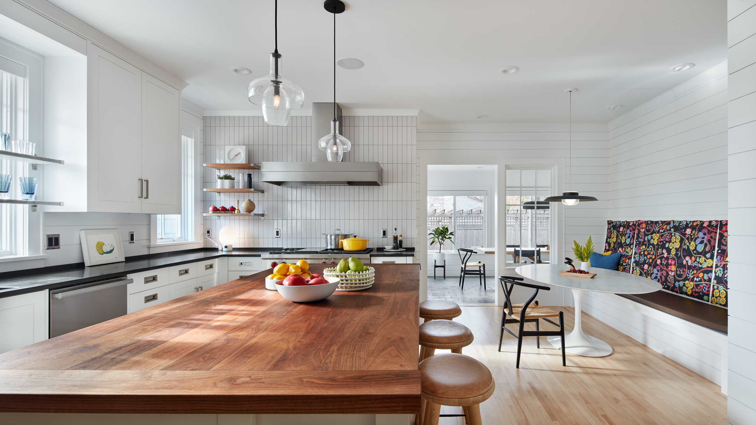 Modern kitchen with wooden island, bar stools, white cabinets, hanging lights, and a bowl of fruit. In the background, a built-in bench with a colorful cushion surrounds a round dining table near large windows, creating a bright and inviting space that captures the spirit of revival in home design.