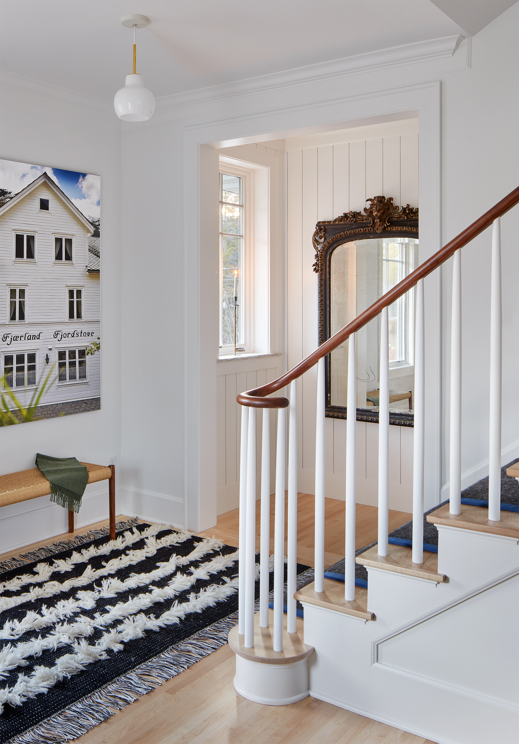 Bright entryway with a large mirror, staircase with wooden handrail, patterned rug, and a bench adorned with a green throw—each element lending a sense of revival to the space. Wall art featuring a white house adds character, while natural light streams in through a window.