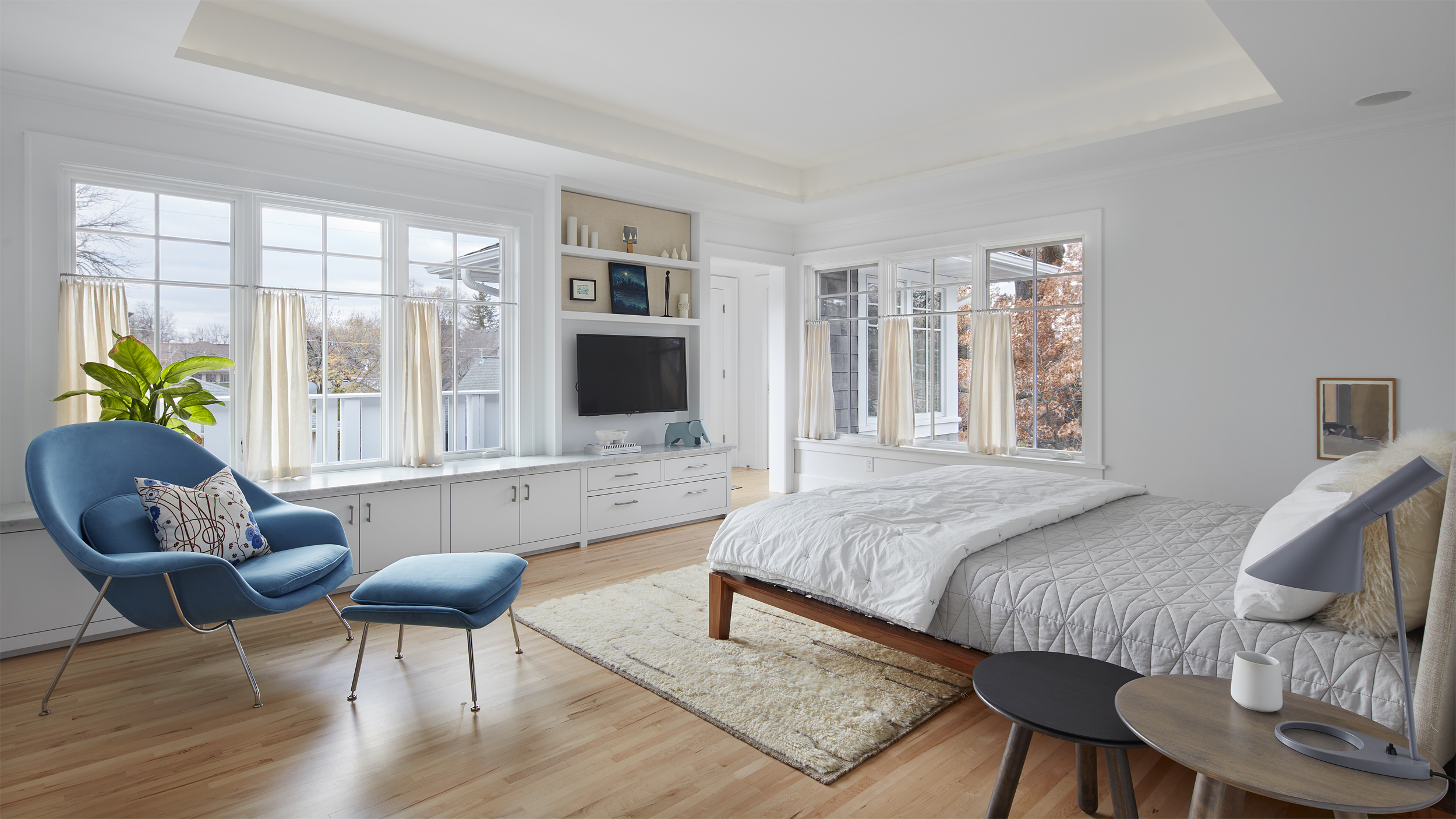 Bright, modern bedroom with large windows, wood floors, and a blue lounge chair offers a sense of revival with its fresh ambiance. The bed is topped with a white quilt, while a TV built into sleek white cabinetry and touches of greenery complement the minimal decor throughout the space.