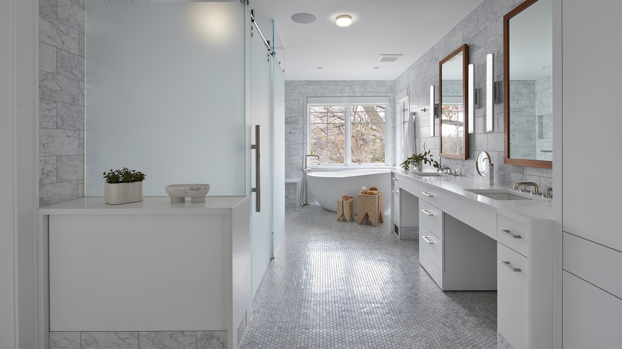 Modern bathroom with white cabinets, double sinks, large mirrors, light gray walls, and a freestanding bathtub by a window evokes a sense of revival. Wooden stools and small plants add to the decor, enhancing the room’s bright, clean, and minimalistic design.