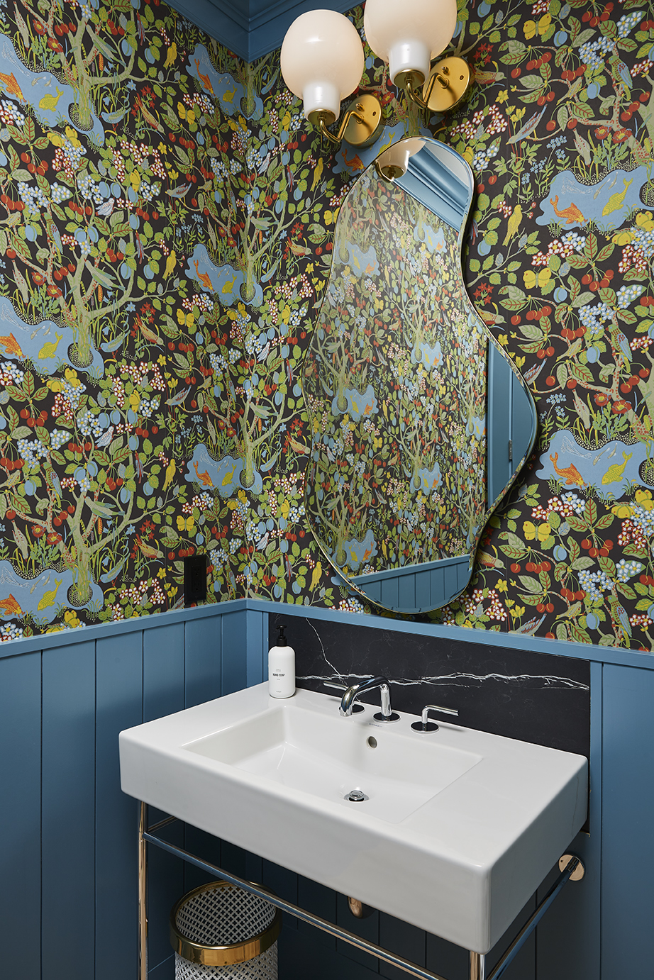 A bathroom with vibrant floral wallpaper exudes a sense of revival, featuring an irregularly shaped mirror, a white sink, blue paneling, a gold sconce with round bulbs, and a small wastebasket underneath the sink.
