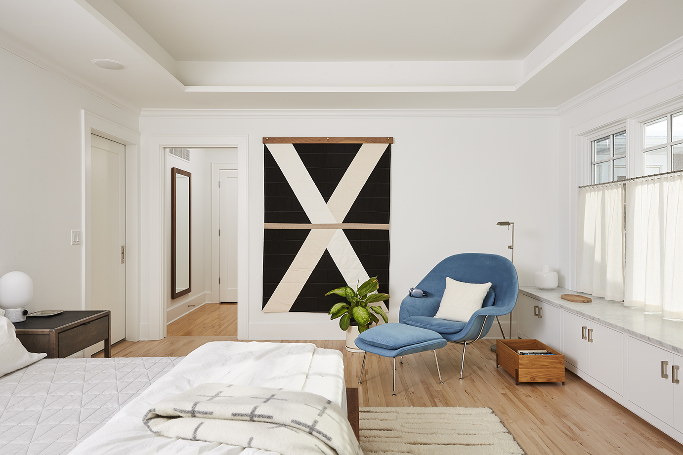 A modern bedroom with a large X artwork on the wall, a blue lounge chair, a potted plant, and a bed with a gray blanket creates an inviting atmosphere. Wood floors and white walls enhance the sense of revival in the space, while natural light from two windows brightens every corner.
