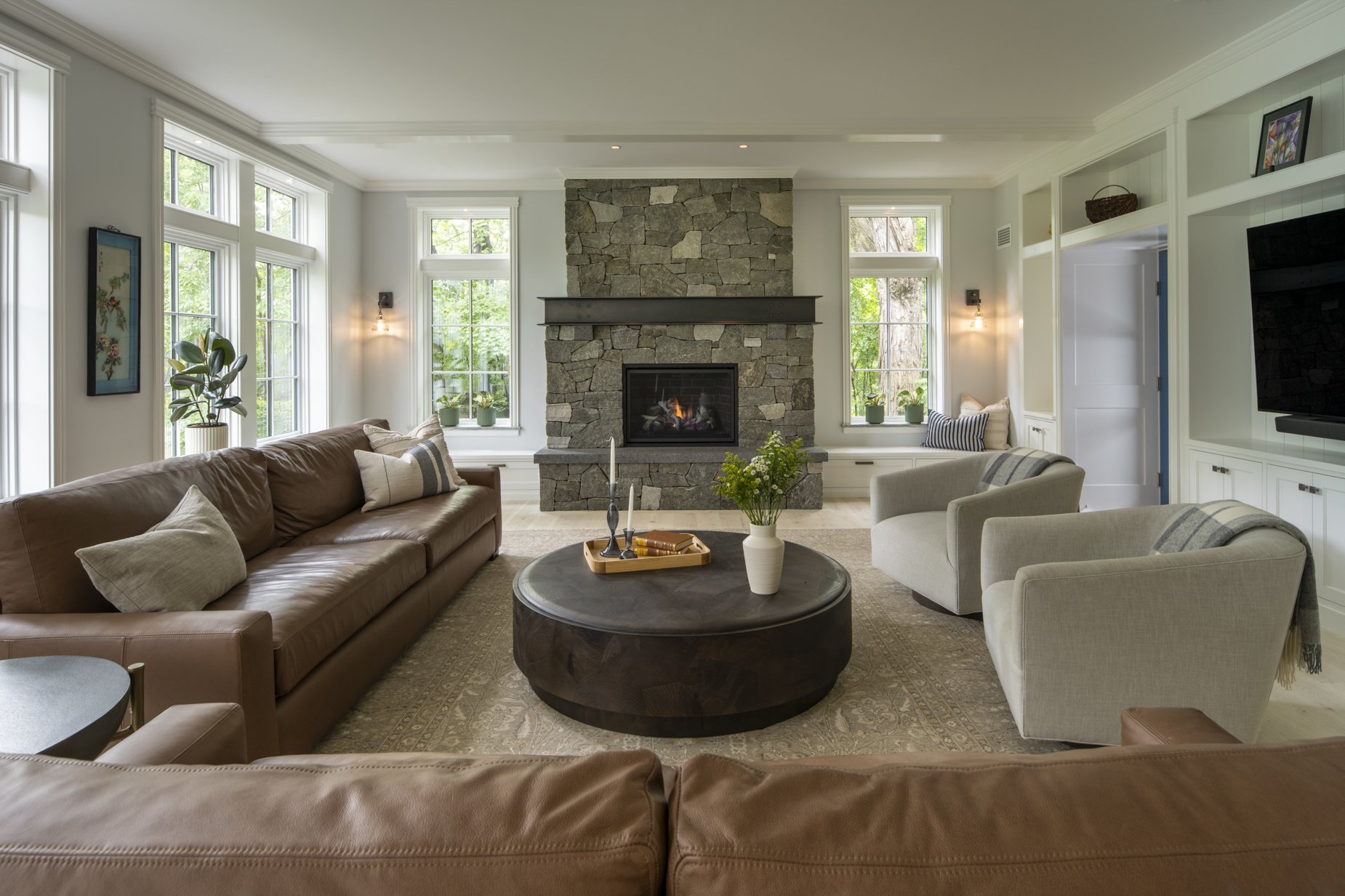 Modern living room with large windows, brown leather sofas, two gray armchairs, a round coffee table, and a stone fireplace with built-in shelves and a TV on the right. Green plants and decor accents are visible throughout.