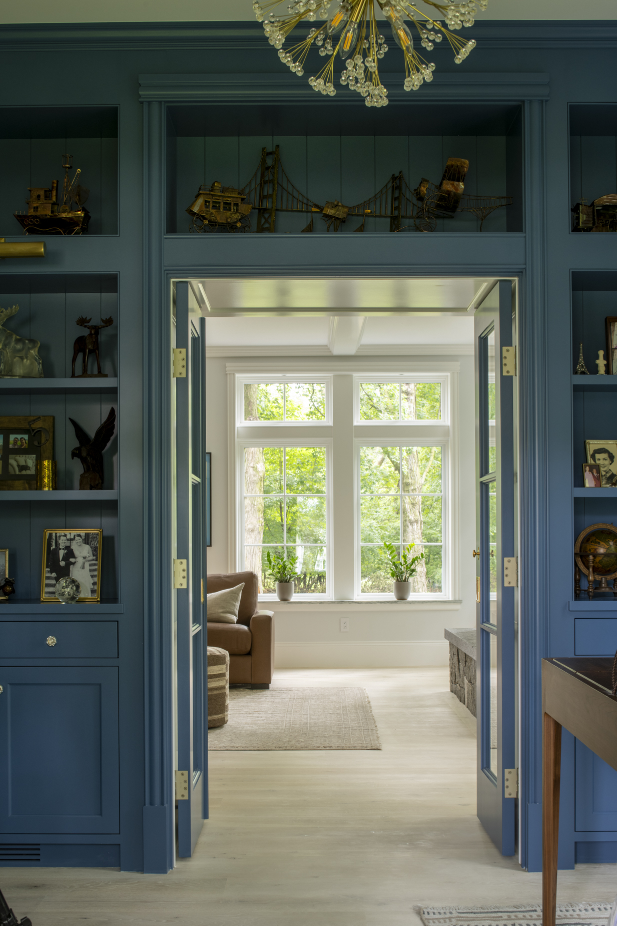 Blue built-in shelves frame open doors leading to a bright sunroom with large windows, brown sofa, beige rug, and green plants. Gold light fixture hangs from the ceiling.