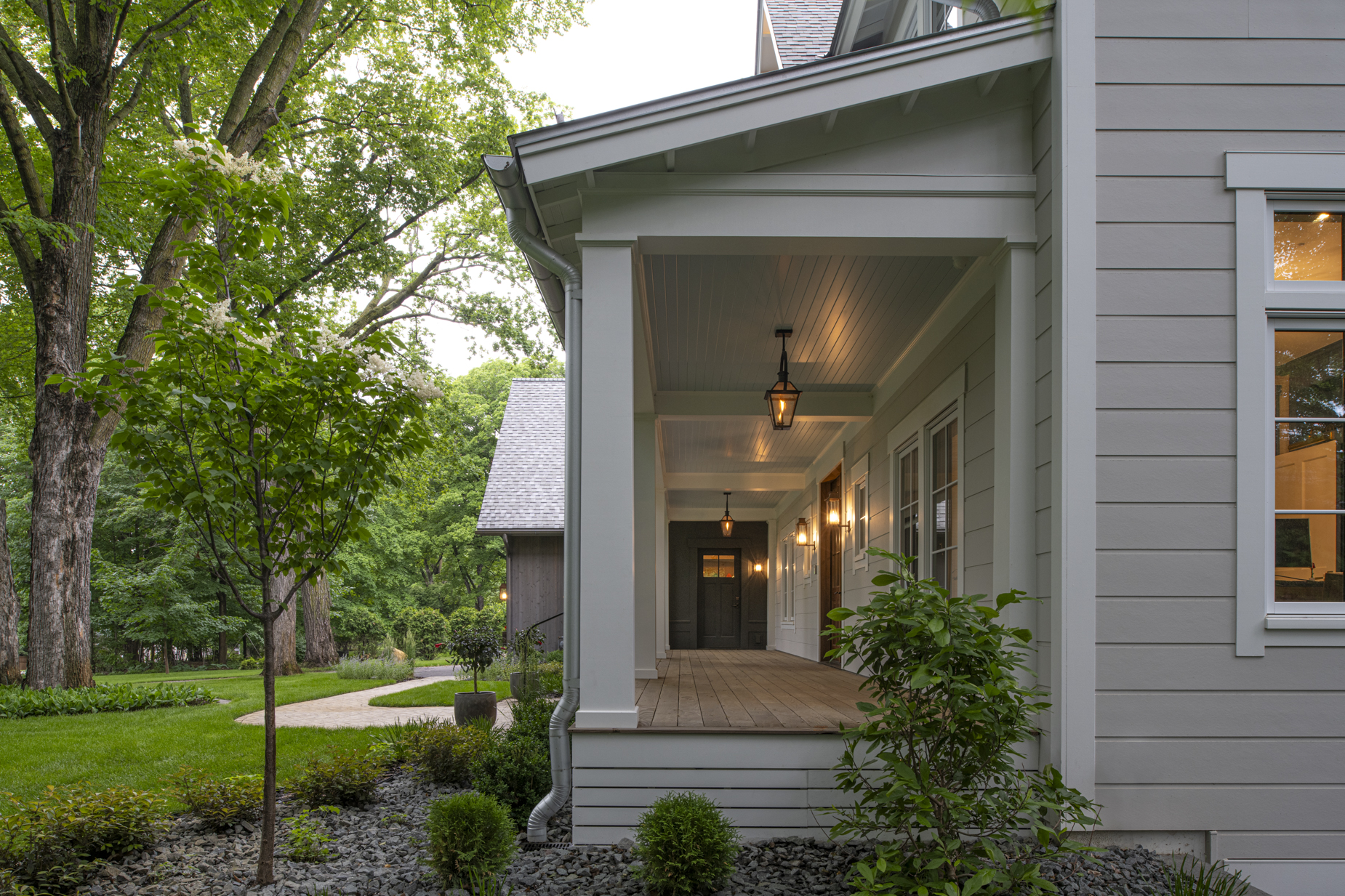 A modern gray house with a covered front porch, warm hanging lights, wood decking, neatly trimmed bushes, a small tree, and a green lawn with large trees in the background.