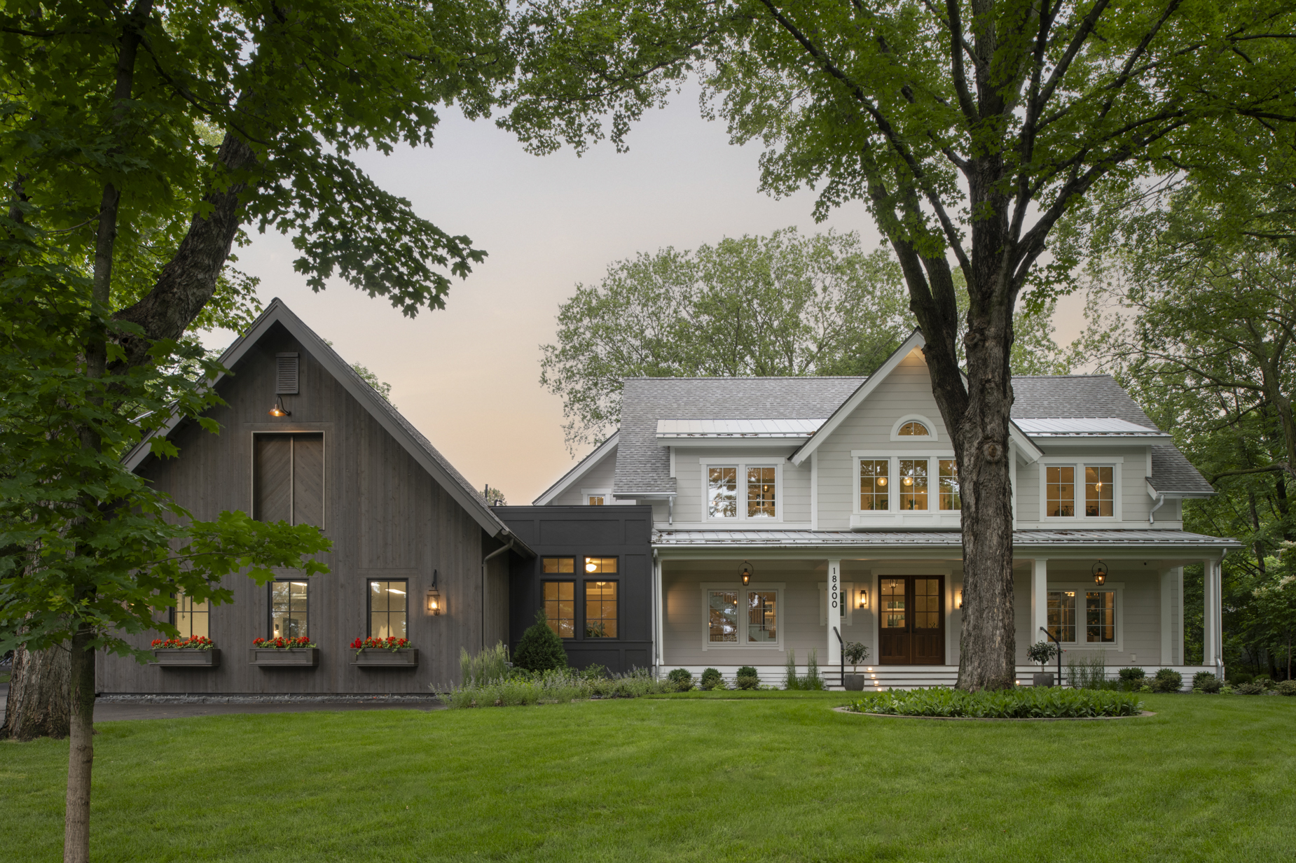 A modern farmhouse with gray and white exteriors, large windows, front porch, flower boxes, and surrounded by green lawn and tall trees at sunset.