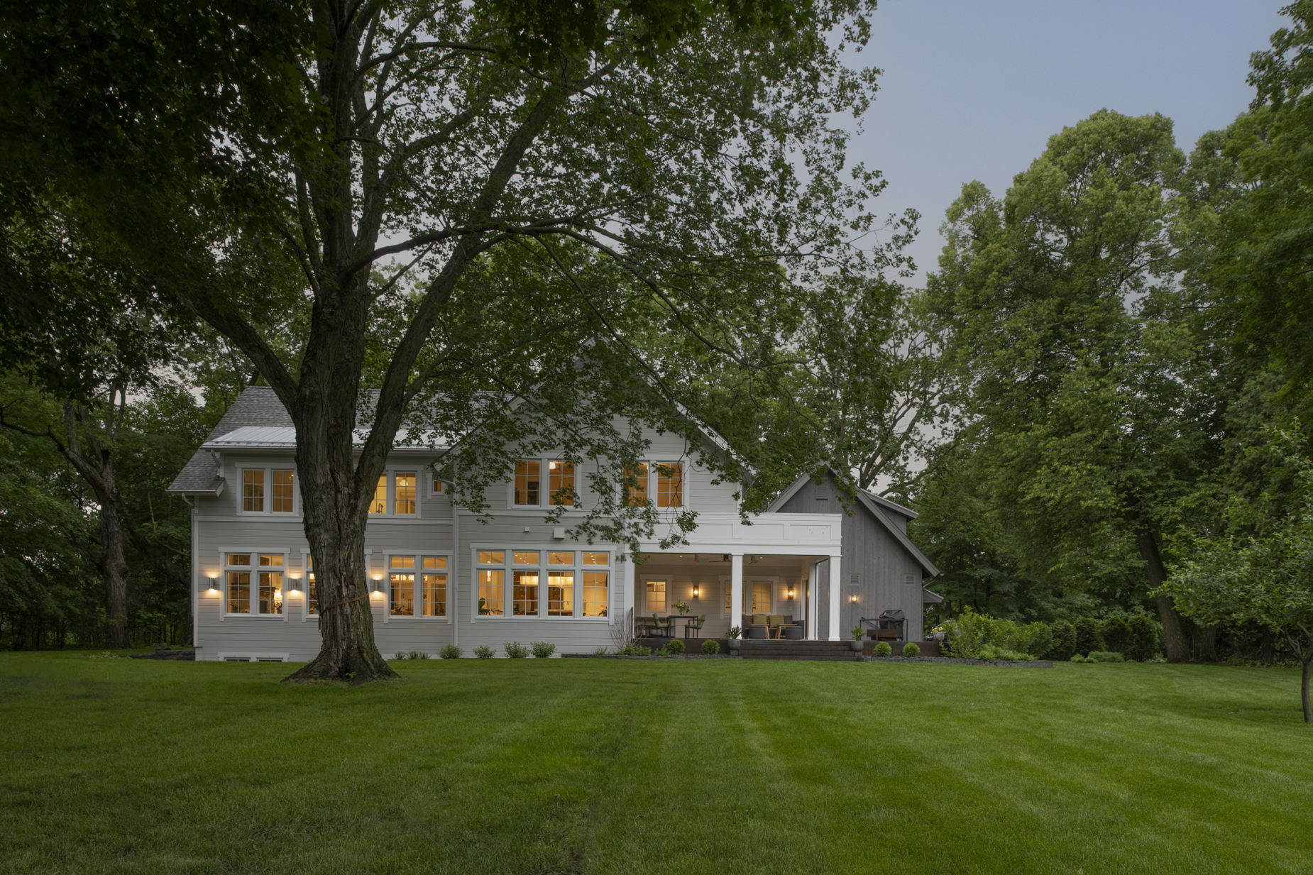 A large white house with glowing windows sits among lush green trees and a well-manicured lawn at dusk.