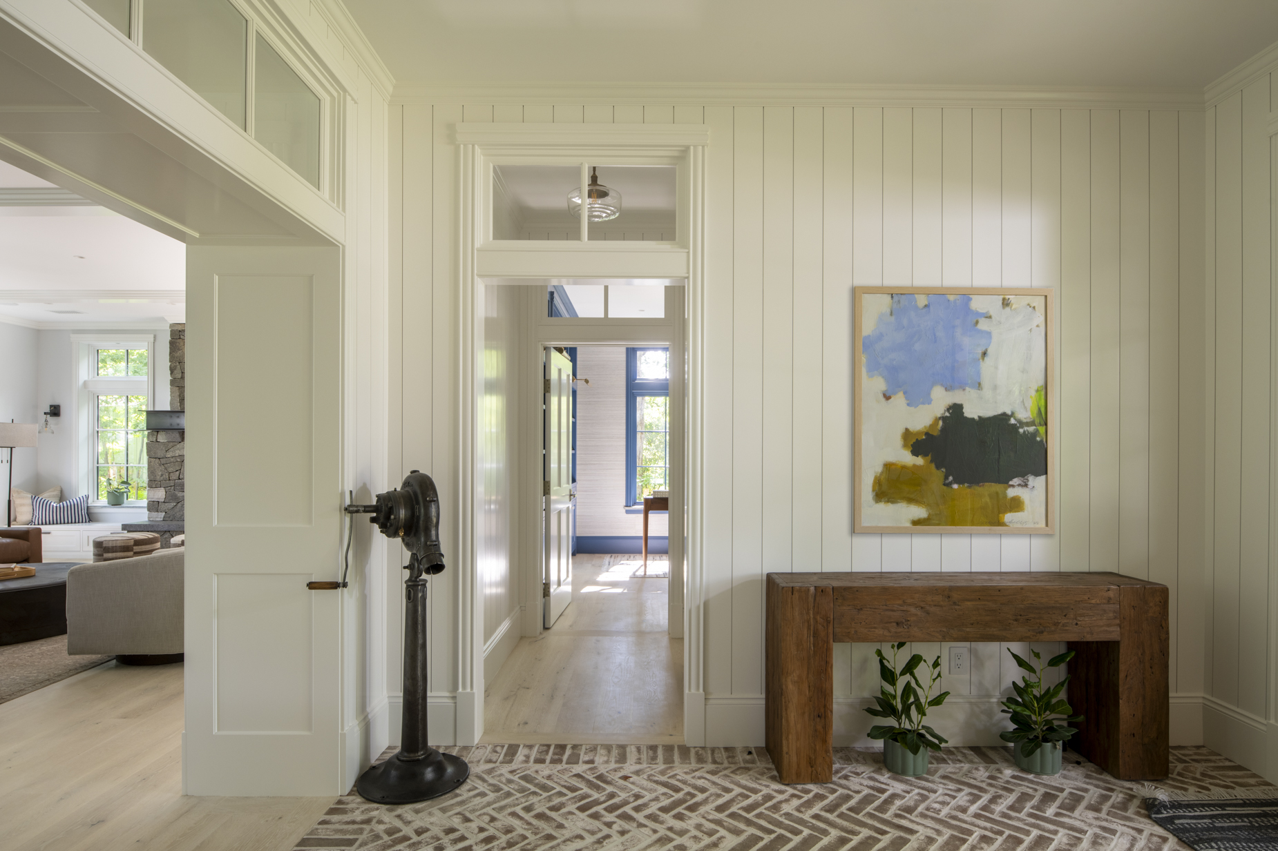 A bright hallway with white panel walls, abstract artwork, a rustic wooden table, two potted plants, and a vintage hand-crank device on a patterned rug. Natural light streams in from adjacent rooms.