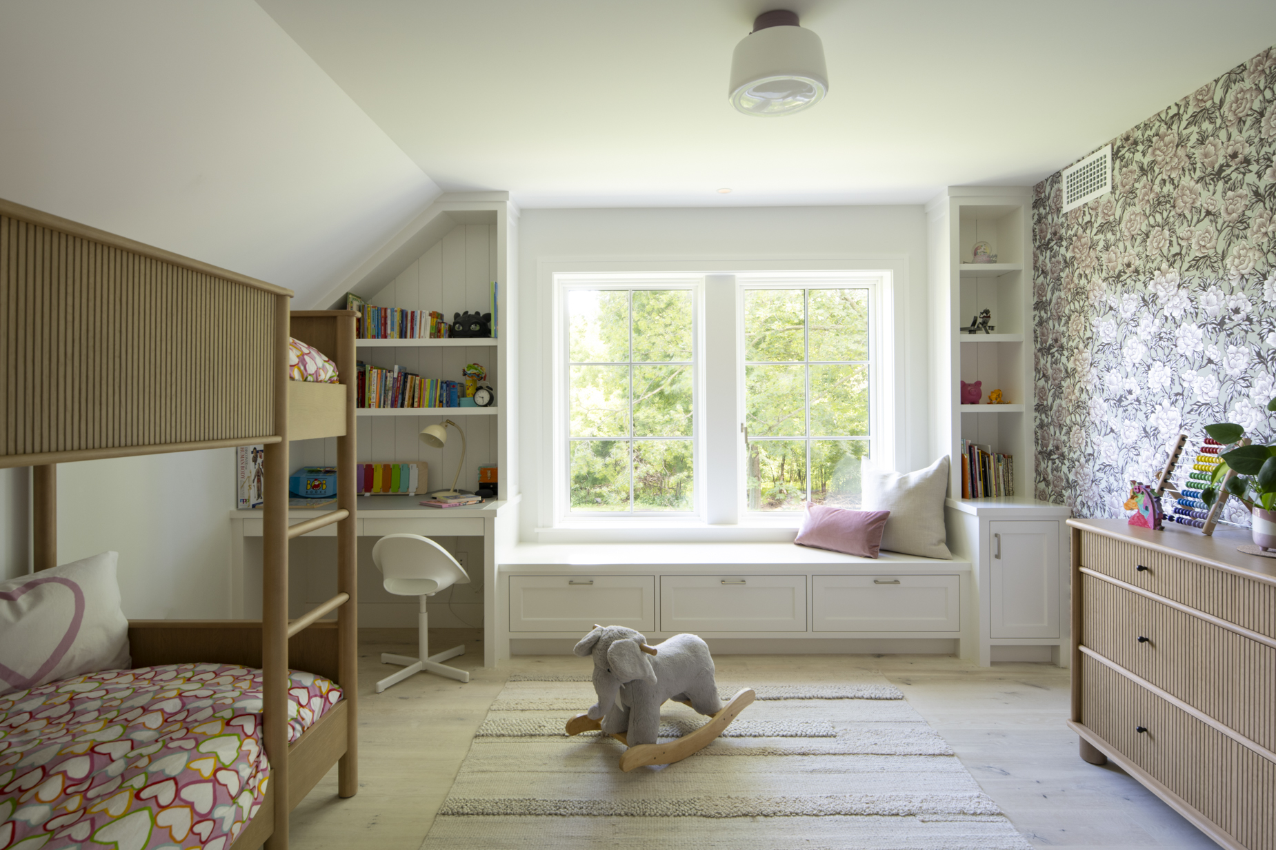 Bright childrens bedroom with bunk beds, a rocking elephant toy, a window seat with cushions, bookshelves, and floral wallpaper. The room features wooden furniture and large windows letting in natural light.