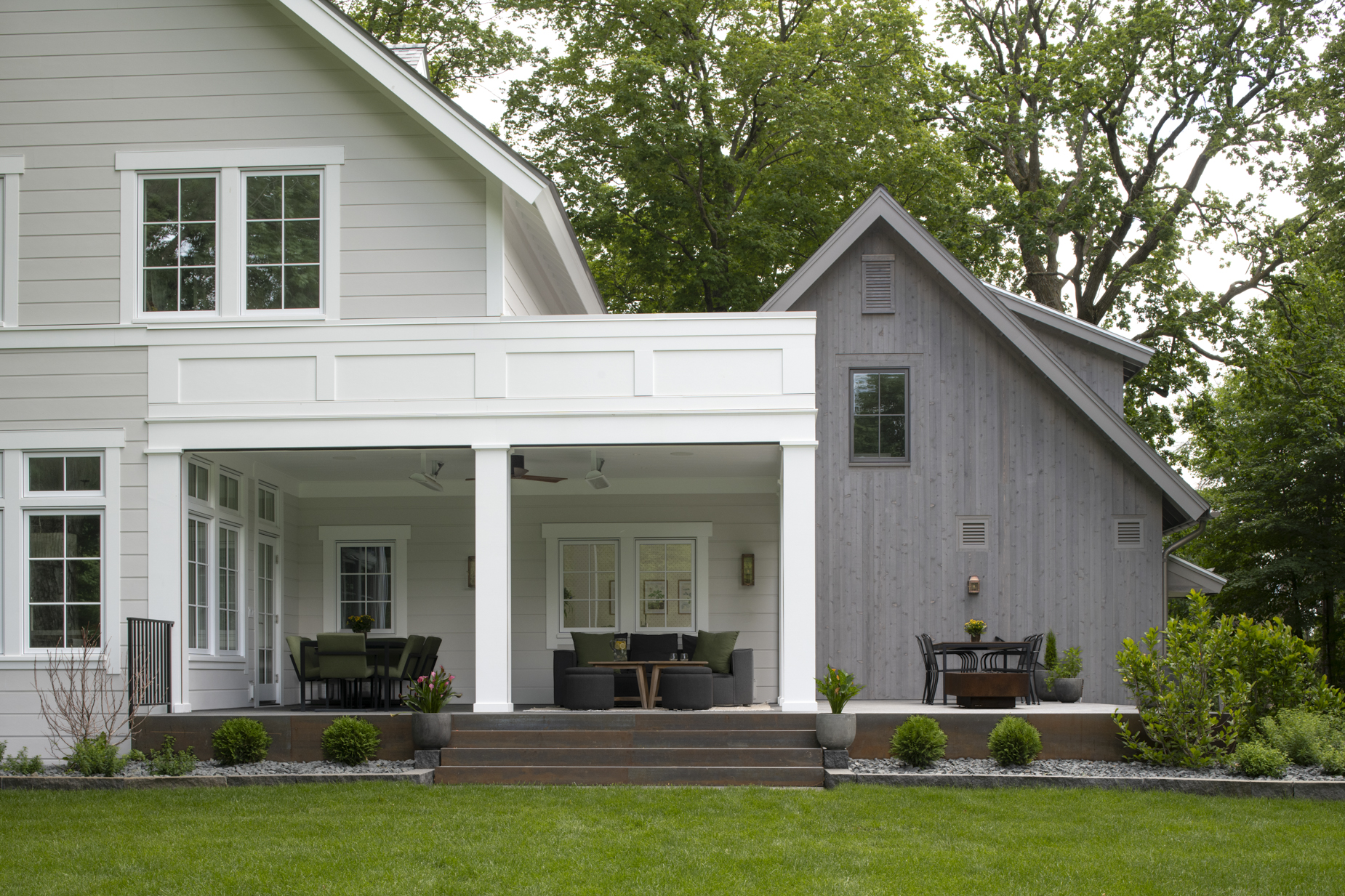 A modern house with a covered patio, outdoor seating areas, and neatly trimmed bushes in front. The exterior features gray siding and large windows, with green lawn and tall trees in the background.