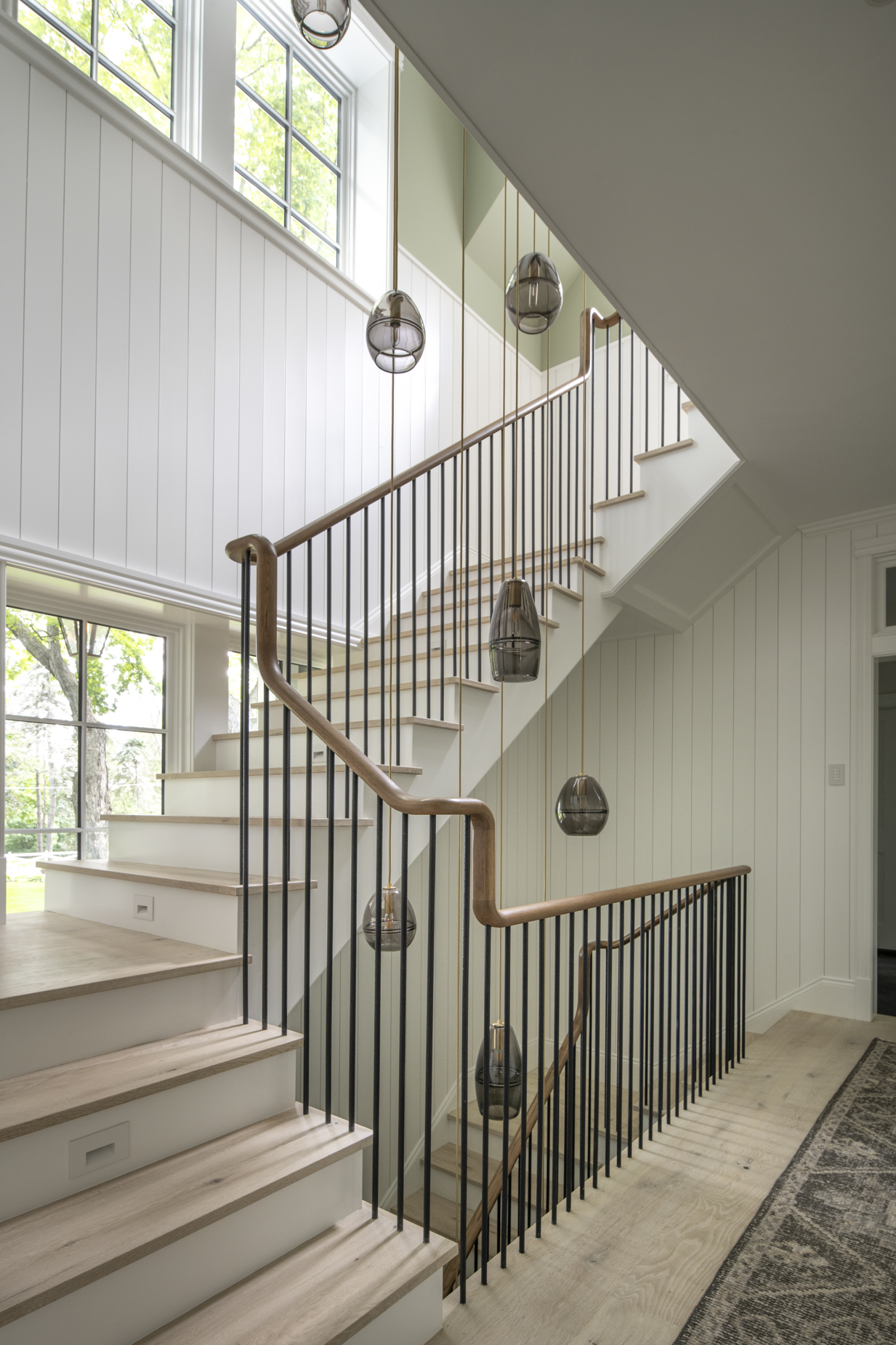 Modern staircase with light wood steps, black vertical railing, wood handrail, and spherical pendant lights. Large windows let in natural light, brightening the white-paneled walls and beige patterned rug below.